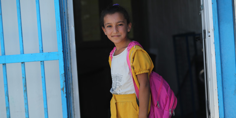 A girl with a white shirt and pink backpack stands in a doorway